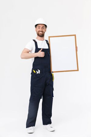 Full length of a confident bearded builder man wearing overalls and hardhat standing isolated over white background, showing blank boardの写真素材
