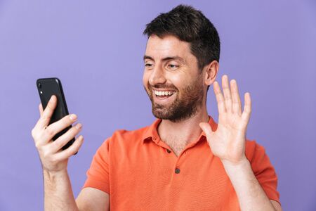 Image of a happy young handsome bearded man posing isolated over violet purple wall background talking by mobile phone waving.の写真素材