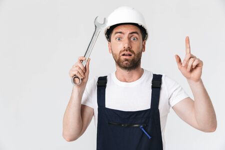 Confused bearded builder man wearing overalls and hardhat standing isolated over white background, showing adjustable wrenchの写真素材