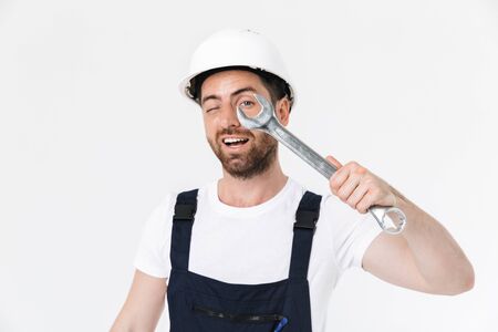 Confident bearded builder man wearing overalls and hardhat standing isolated over white background,...