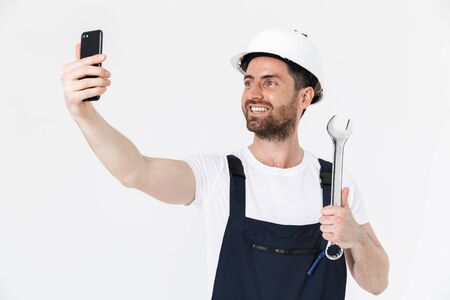 Confident bearded builder man wearing overalls and hardhat standing isolated over white background, showing adjustable wrenchの写真素材