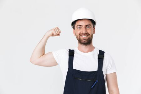 Confident bearded builder man wearing overalls and hardhat standing isolated over white background, flexing musclesの写真素材