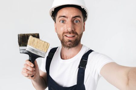 Confident bearded builder man wearing overalls and hardhat standing isolated over white background, taking a selfieの写真素材