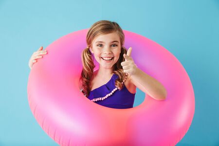 Cheerful little girl wearing swimsuit standing isolated over blue background, playing with...