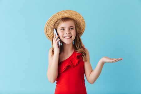 Cheerful little girl wearing swimsuit and summer hat standing isolated over blue background, talking...