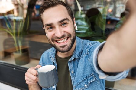 Photo of handsome young man wearing earpods smiling and drinking cup of coffee while taking selfie...