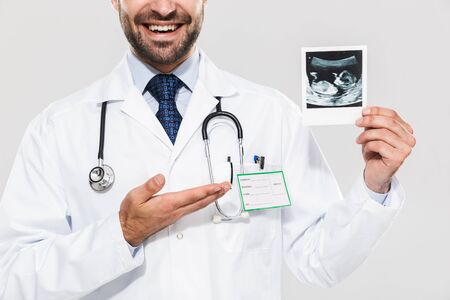 Portrait of cheerful young medical doctor working in hospital and holding ultrasound scan of baby isolated over white backgroundの写真素材