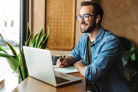Photo of smiling bearded man wearing eyeglasses writing notes and using laptop while working in cafe indoorsの写真素材