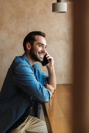 Photo of masculine satisfied man wearing denim shirt smiling and talking on cellphone while sitting on chair in cafe indoorsの写真素材