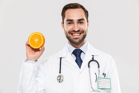 Attractive young cheerful male doctor wearing unifrom standing isolated over white background, showing sliced orangeの写真素材