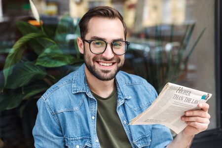 Photo of cheerful young man wearing eyeglasses smiling and reading newspaper while sitting in city cafe outdoorsの写真素材