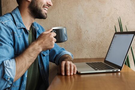 Image of handsome bearded man wearing denim shirt drinking coffee and using laptop while sitting at...