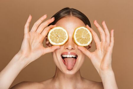 Image of a beautiful happy young amazing woman posing isolated over brown chocolate background wall holding lemon covering eyes.の写真素材