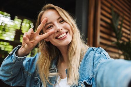 Image of a happy young pretty blonde woman in cafe take a selfie by camera showing peace.の写真素材