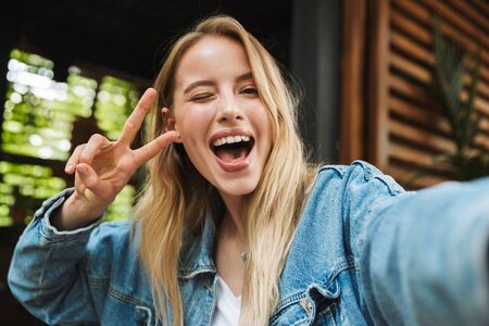 Image of amazing excited happy young woman posing outdoors in cafe take a selfie by camera showing peace gesture.の写真素材