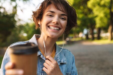 Attractive cheerful young girl wearing casual outfit spending time outdoors at the park, listening to music with earphones, showing takeaway coffee cupの写真素材