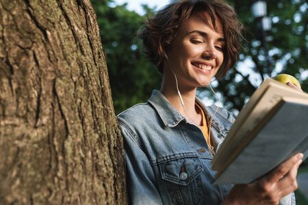 Attractive smiling young girl wearing casual outfit spending time outdoors at the park, leaning on a...