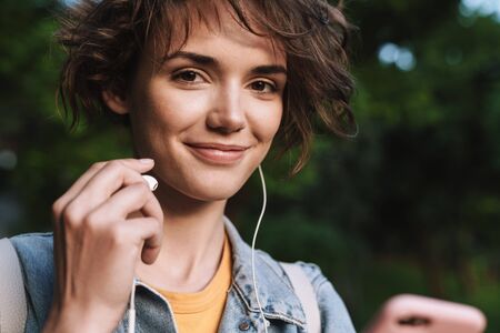 Attractive happy young girl wearing casual outfit spending time outdoors at the park, listening to music with earphones, holding mobile phoneの写真素材