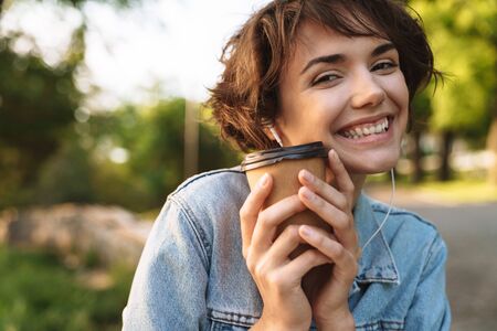 Image of delighted caucasian woman 20s wearing denim jacket smiling while drinking takeaway coffee...