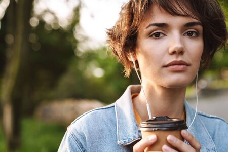 Image of thoughtful caucasian woman 20s wearing denim jacket drinking takeaway coffee and using...