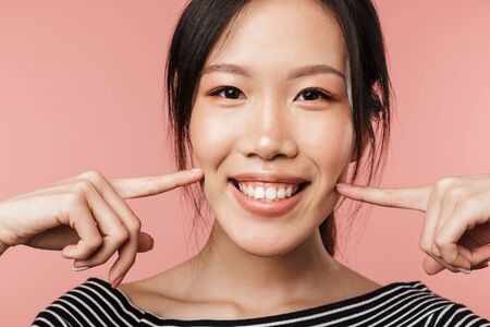 Close up of a smiling cute asian woman standing isolated over pink background, grimacingの写真素材