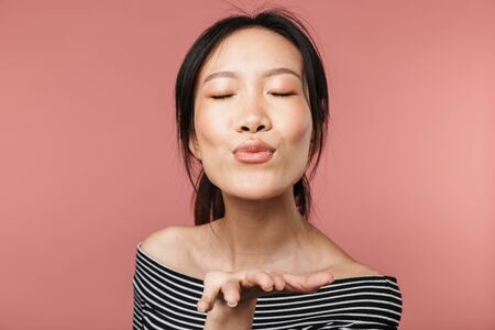 Portrait of a lovely young asian woman standing isolated over pink background, sending kissの写真素材