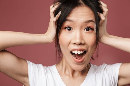 Portrait of astonished asian woman dressed in basic wear wondering and looking at camera while grabbing head isolated over red background in studioの写真素材