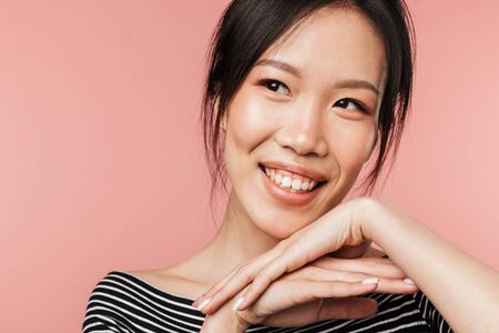 Photo closeup of cheery asian woman dressed in basic wear smiling and looking aside isolated over red background in studioの写真素材