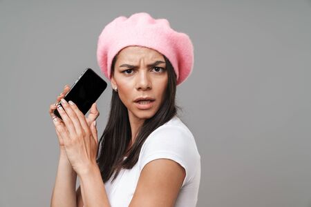 Photo closeup of puzzled pretty woman in basic t-shirt and beret frowning while holding smartphone isolated over gray backgroundの写真素材