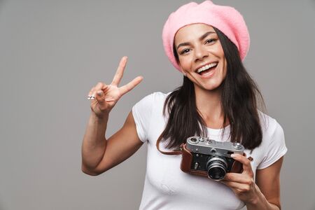Photo closeup of cheery pretty tourist woman in beret laughing and showing peace sign while holding retro camera isolated over gray backgroundの写真素材