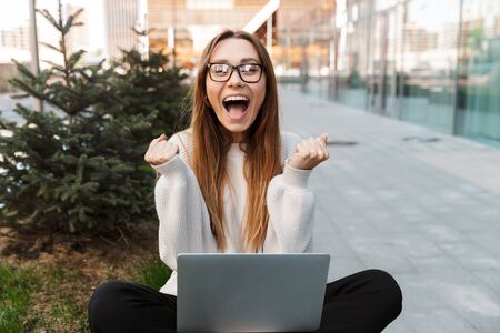 Image of a beautiful excited shocked young happy business woman posing sitting outdoors near business center wearing eyeglasses using laptop computer make winner gesture.の写真素材