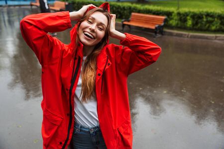 Image of attractive young woman 20s in colorful raincoat having fun while walking through green park under rainの写真素材