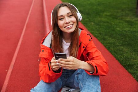 Cheerful attractive young girl wearing raincoat spending lovely time outdoors, listening to music...