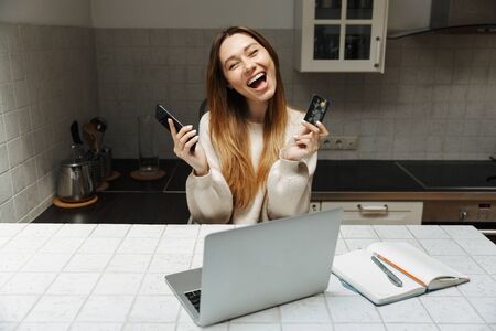 Attractive young girl sitting at the kitchen at home with laptop computer, using mobile phone and credit cardの写真素材