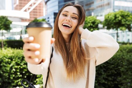 Beautiful young cheerful woman walking outdoors at the city street, drinking takeaway coffeeの写真素材