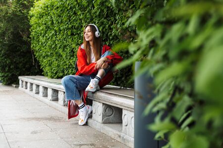 Cheerful attractive young girl wearing raincoat spending lovely time outdoors, listening to music with headphones, sitting on a benchの写真素材