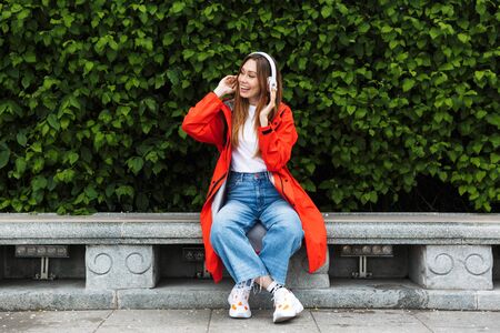 Cheerful attractive young girl wearing raincoat spending lovely time outdoors, listening to music with headphones, sitting on a benchの写真素材