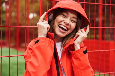 Cheerful attractive young girl wearing raincoat spending lovely time outdoors, listening to music...