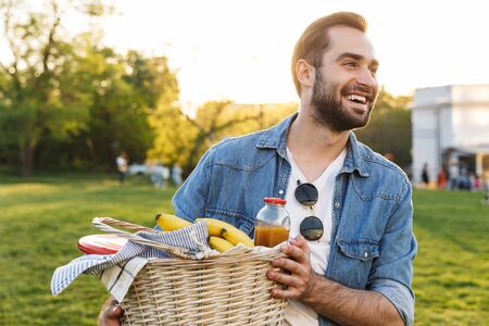 Happy young bearded man walking at the park, carrying picnic basket full of foodの写真素材