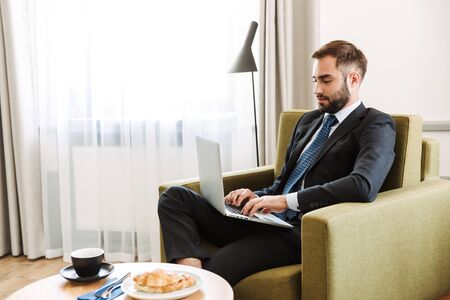 Attractive young businessman wearing suit sitting in a chair at the hotel room, working on laptop computer while having breakfastの写真素材