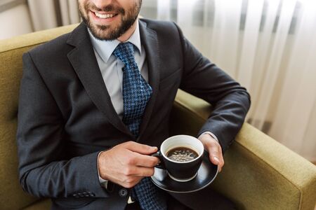 Cropped image of an attractive young businessman wearing suit sitting in a chair at the hotel room,...
