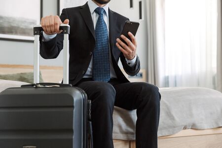 Cropped image of a young businessman wearing suit sitting at the hotel room, using mobile phone...