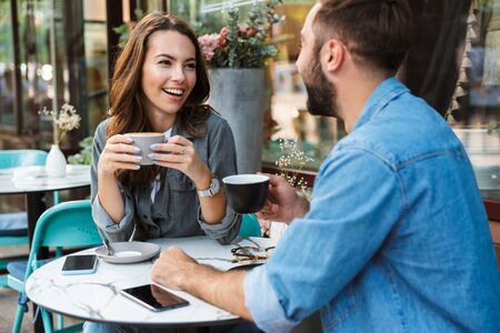 Attractive young couple in love having lunch while sitting at the cafe table outdoors, drinking coffee, talkingの写真素材