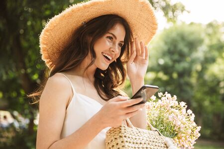 Image of excited happy young pretty woman outoors in green park walking in hat using mobile phone.