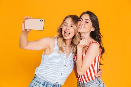 Two cheerful young girls wearing summer outfit, standing isolated over yellow background, taking a selfieの写真素材
