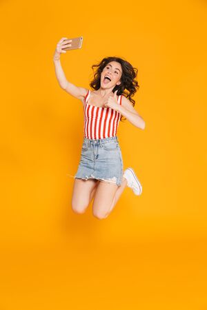 Portrait of excited brunette woman in denim skirt jumping and showing thumb up while taking selfie on cellphone isolated over yellow backgroundの写真素材