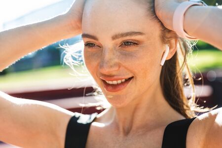 Photo closeup of joyful sportswoman 20s wearing tracksuit and earpods touching her hair during workout in city parkの写真素材