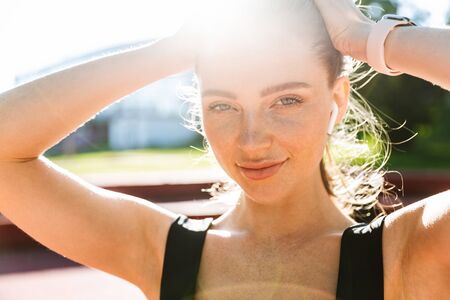 Photo closeup of sunlit sportswoman 20s wearing tracksuit and earpods smiling and looking at camera...