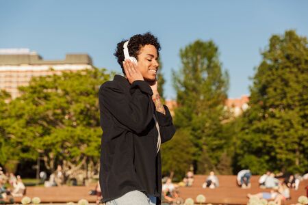 Portrait closeup of smiling african american woman with curly afro hairstyle listening to music on headphones while walking on city parkの写真素材