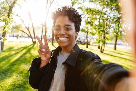 Photo of charming african american woman with curly hair showing peace sign and taking selfie on...